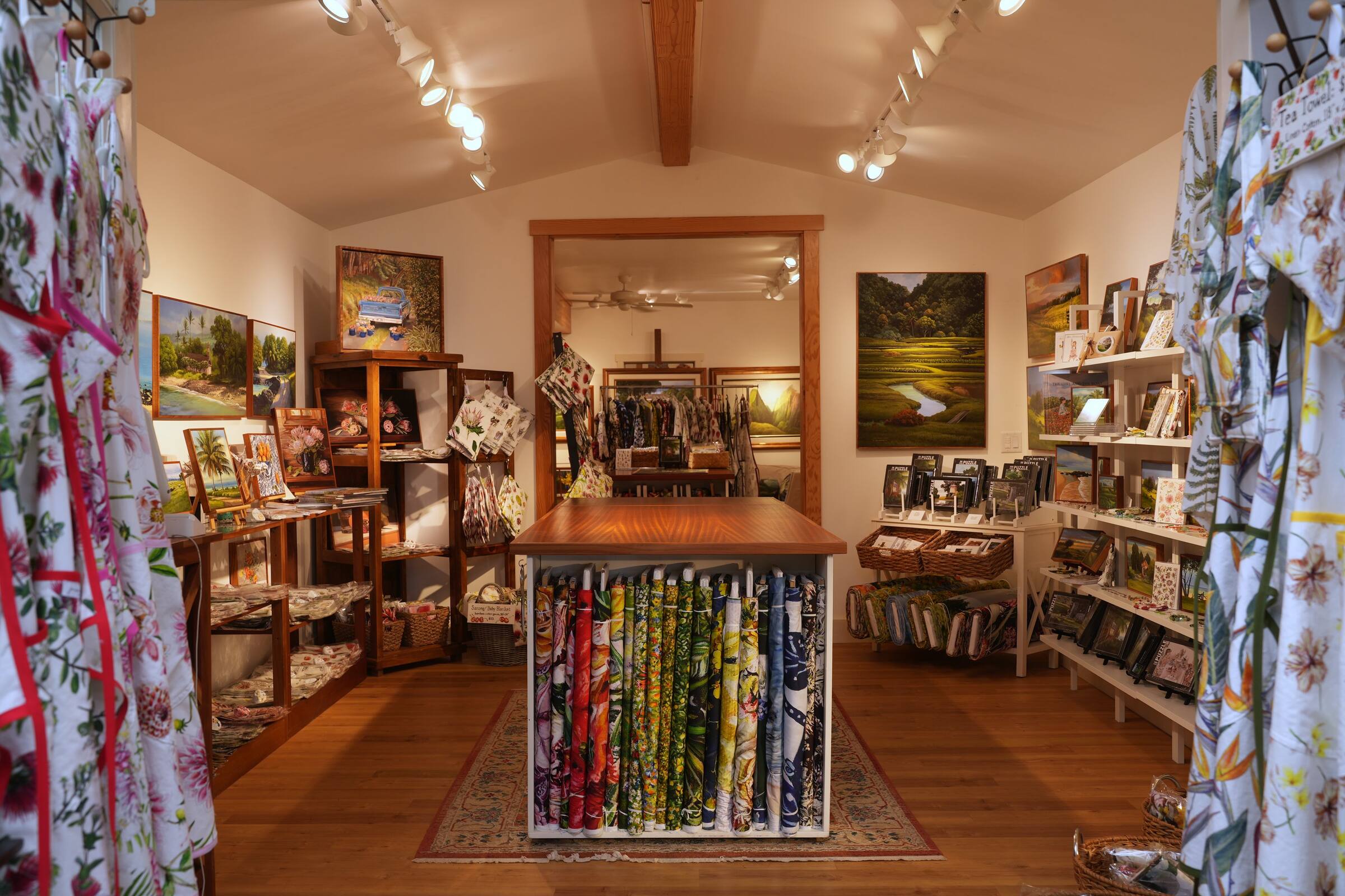 The gift shop from its doorway, fabrics stacked in the middle and paintings on every wall